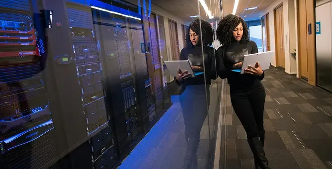 A woman works on a laptop in a server room, her reflection visible.