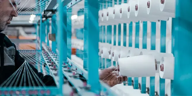A worker adjusts white thread spools on a blue textile manufacturing machine.