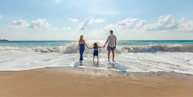 Family holding hands on a sandy beach with ocean waves under a sunny sky.