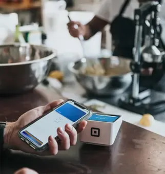 A hand uses a smartphone for mobile payment at a small square terminal.