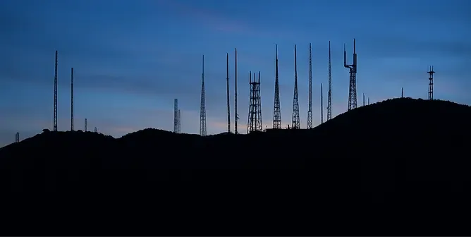 Silhouette of numerous communication towers on a dark hilltop against a twilight blue sky.