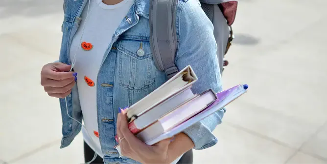 A student wearing a denim jacket walks with a backpack and holds binders.