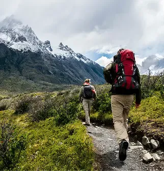 Two hikers on a dirt path with backpacks in front of large snow-capped mountains.