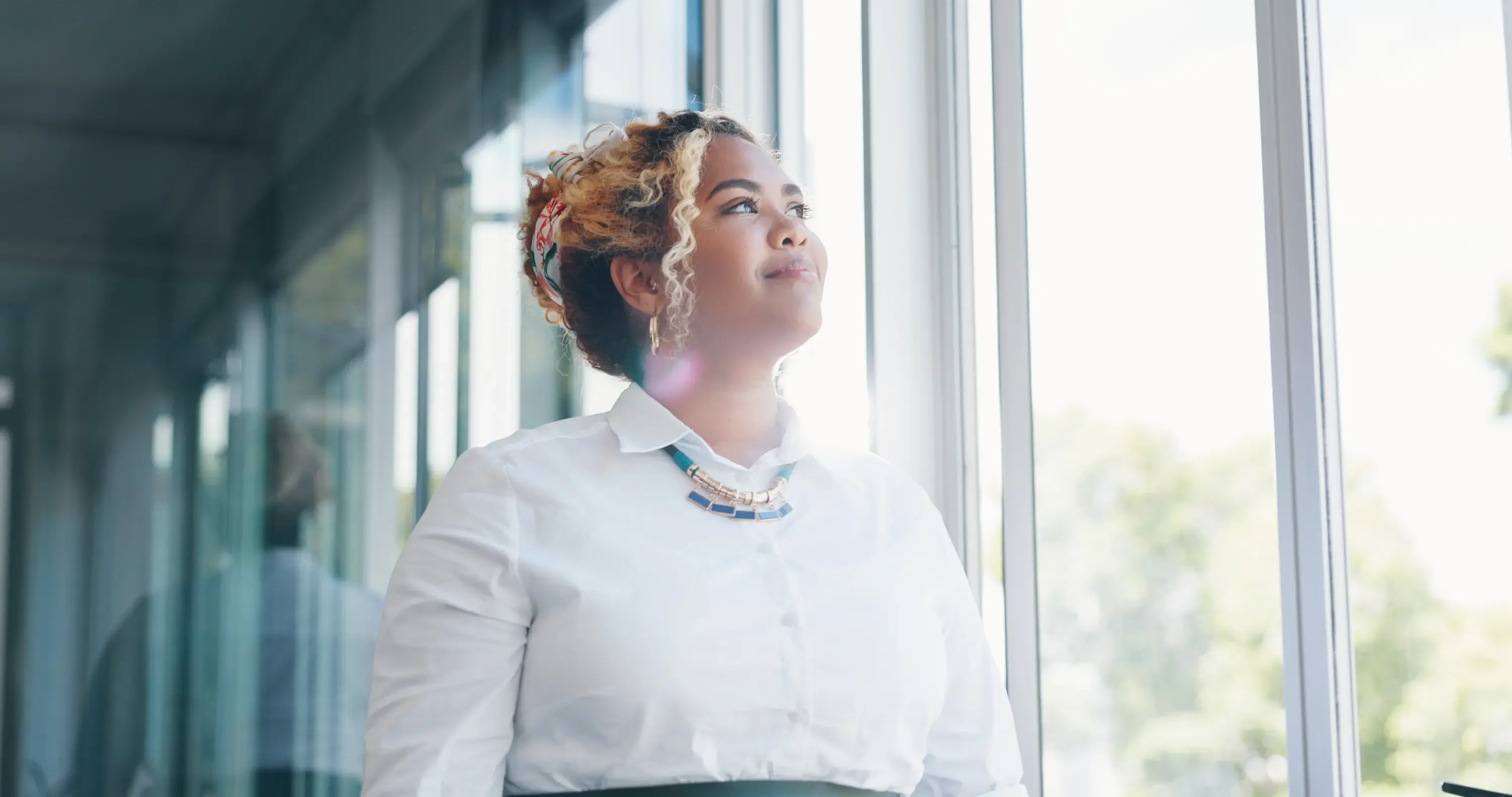 Thoughtful woman with curly hair looking out a large window in a modern building.