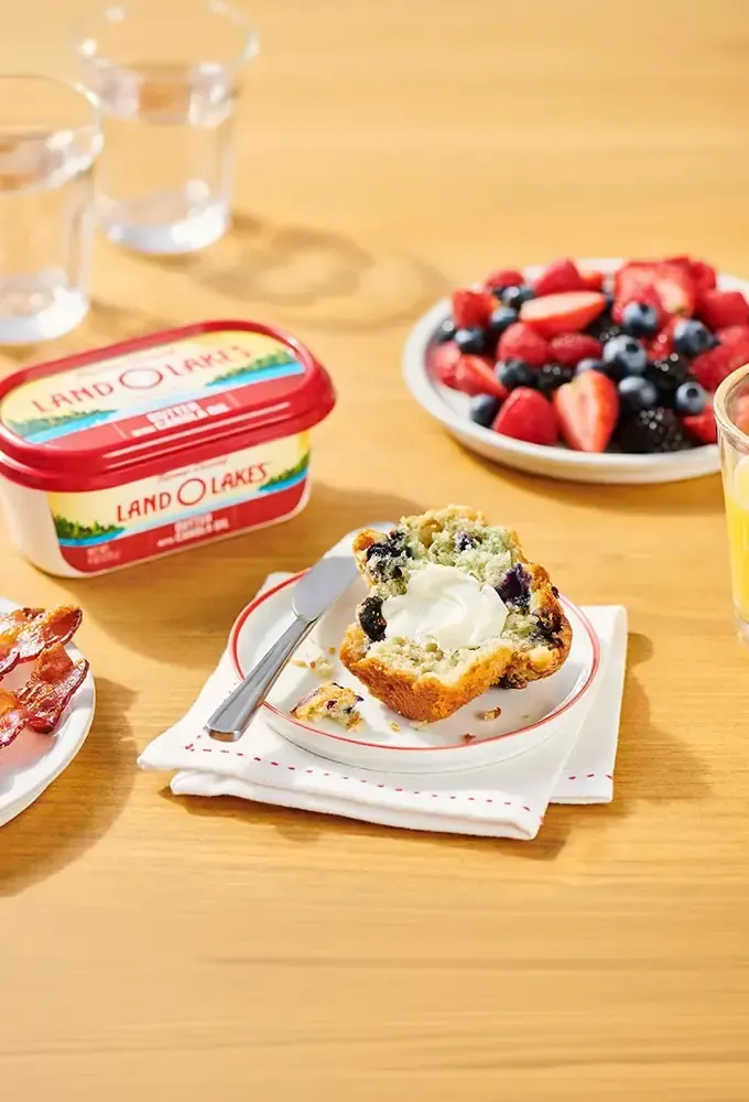 Blueberry muffin with cream cheese, alongside fresh berries, bacon, and a cream cheese container.