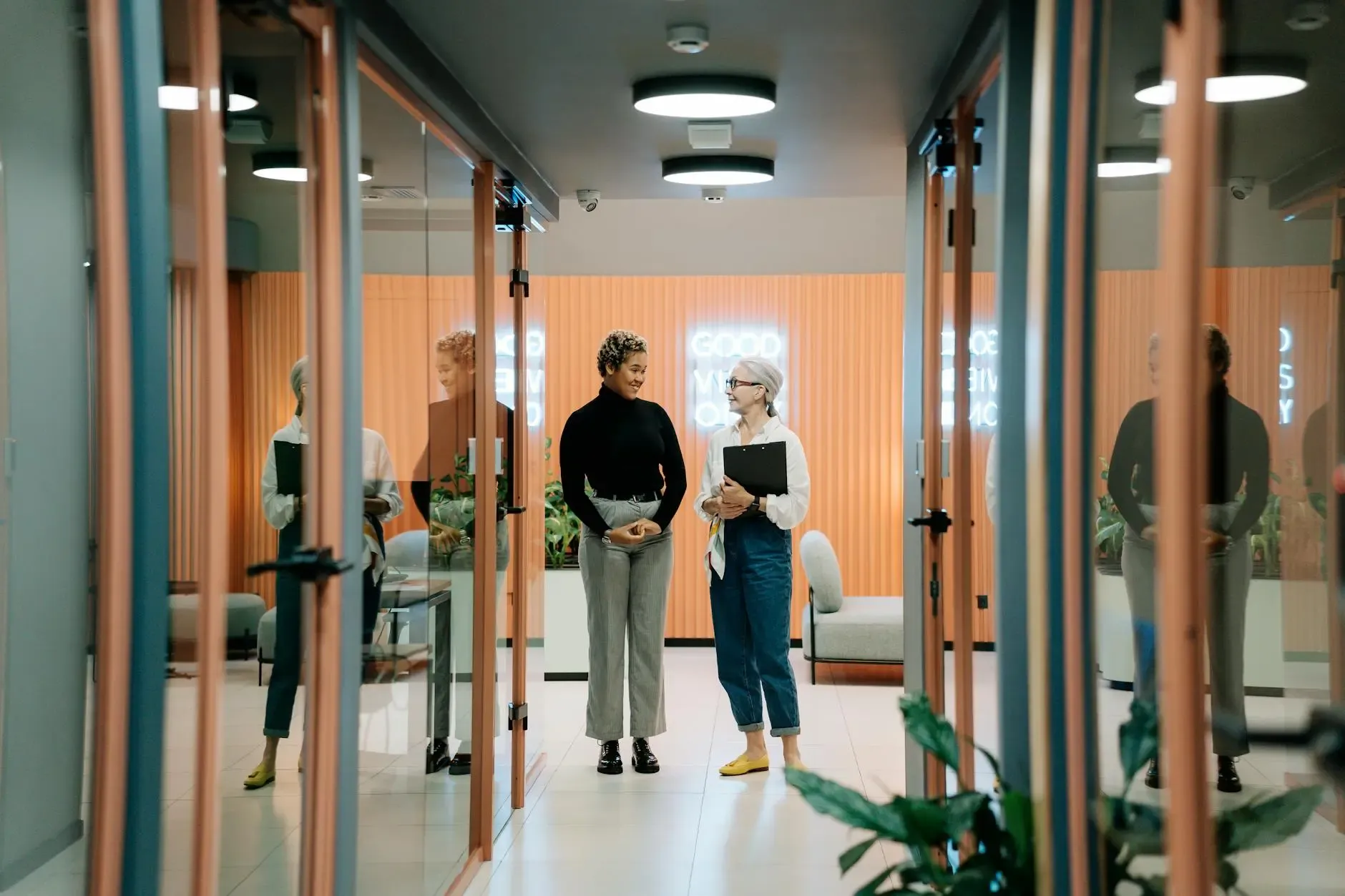 Two women converse in a bright, modern office hallway with glass doors.