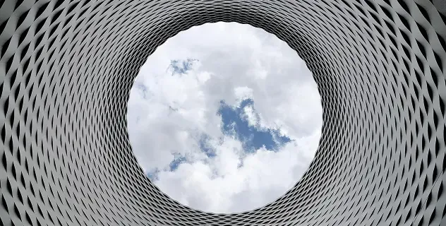 View looking up through a circular, gridded tunnel towards a bright blue sky with white clouds.