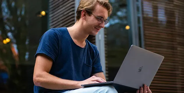 Smiling young man in glasses focused on his laptop screen outdoors.