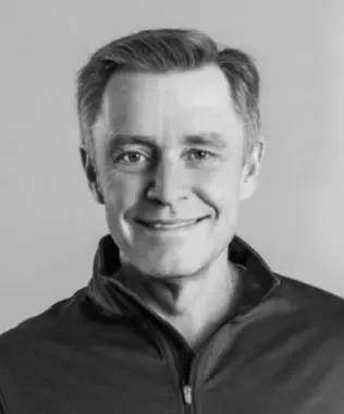 Black and white headshot of a smiling man with short brown hair in a zip-up jacket.