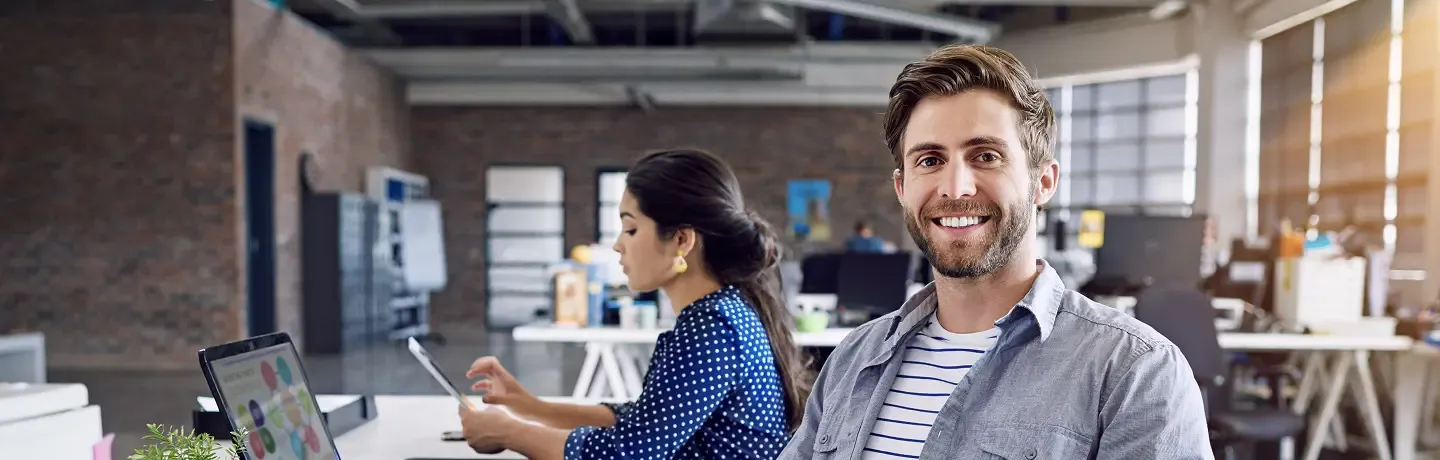 Smiling man with a beard in a modern office, a coworker works in background.
