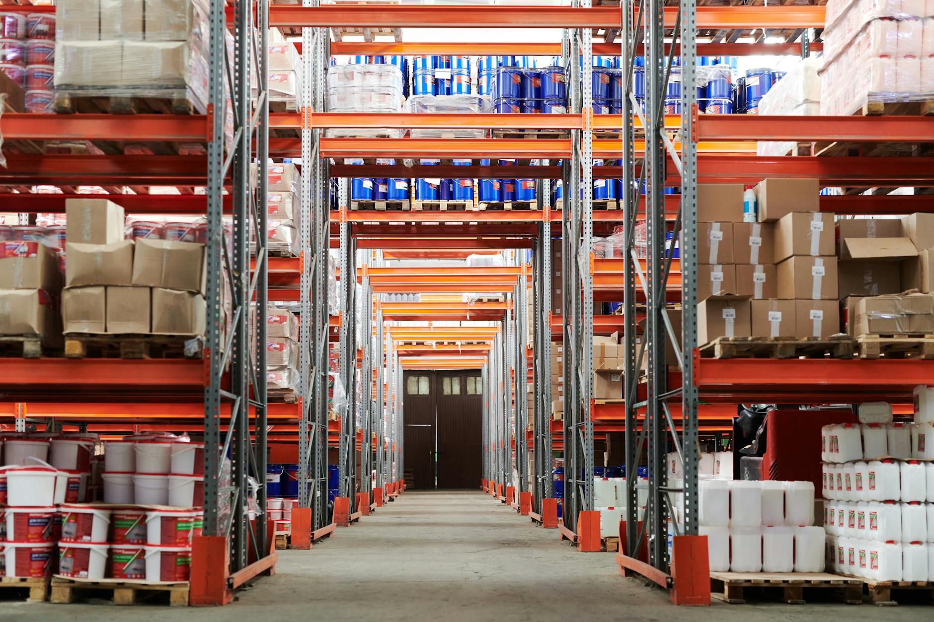 Long aisle in a large industrial warehouse with tall orange racks filled with boxes.