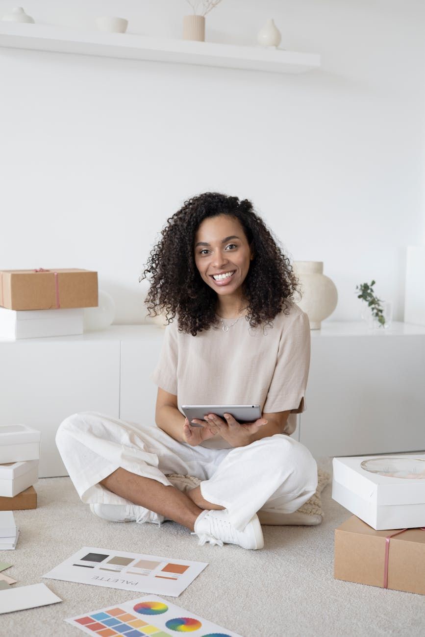 Smiling woman sitting cross-legged on the floor, holding a tablet amidst packing supplies.