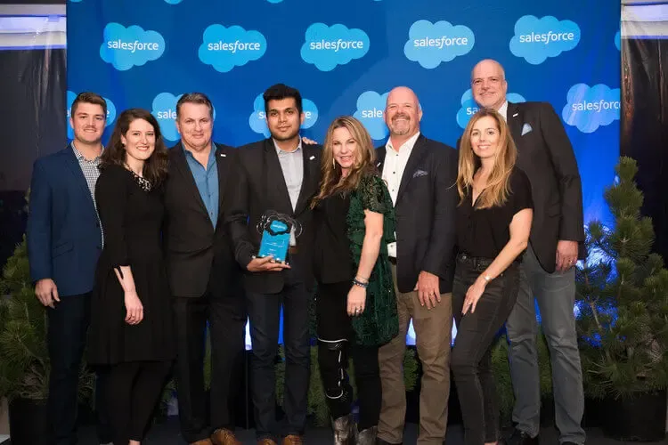 Group of eight diverse people proudly holding a Salesforce award on stage.