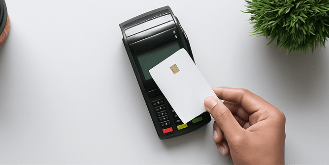 Hand tapping a white payment card on a black point-of-sale terminal for contactless payment.