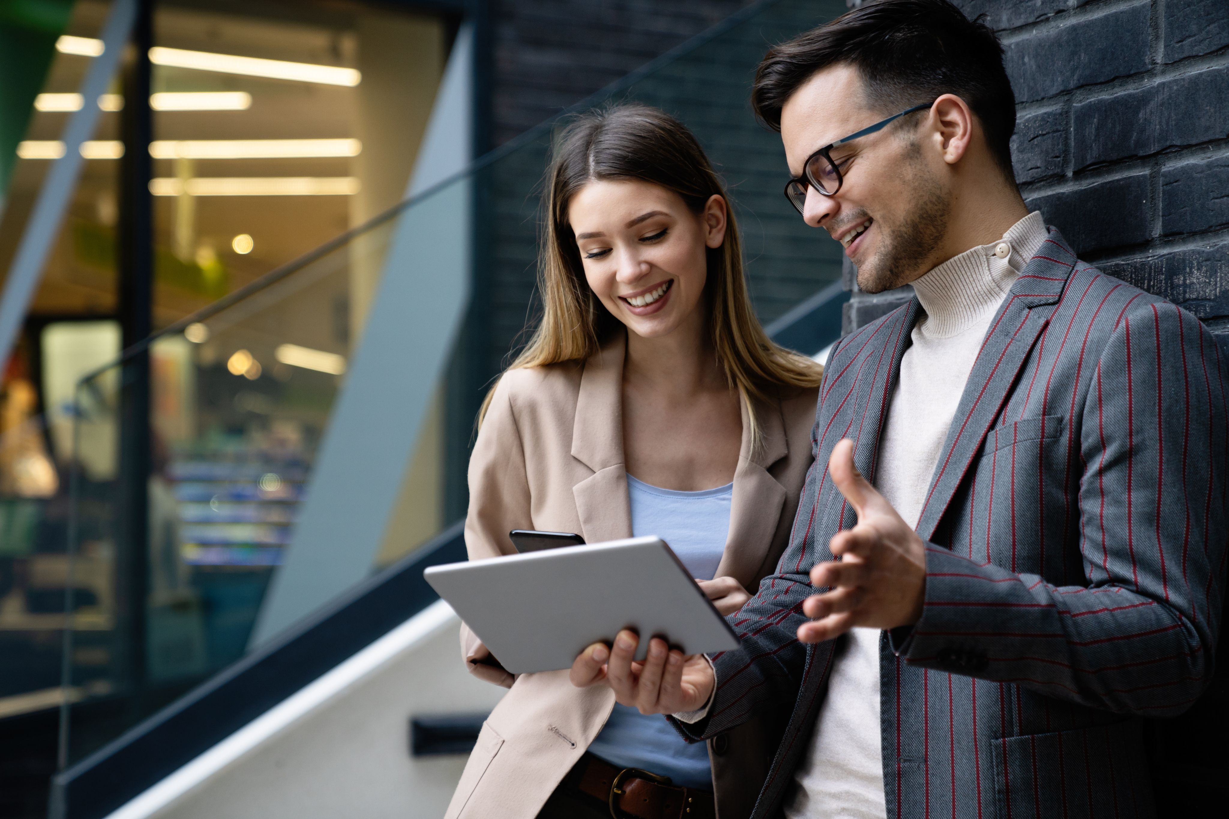 Two smiling professionals review content on a tablet outside a modern office building.