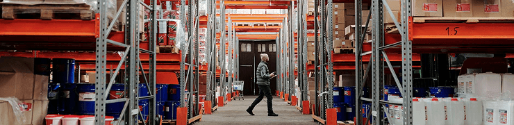 A man walks down a long aisle between tall, fully stocked warehouse shelves.