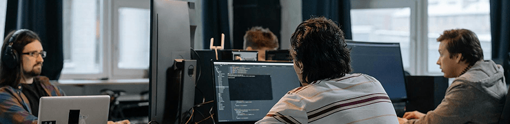 Three men are working intently on computers in a modern office environment with large windows.