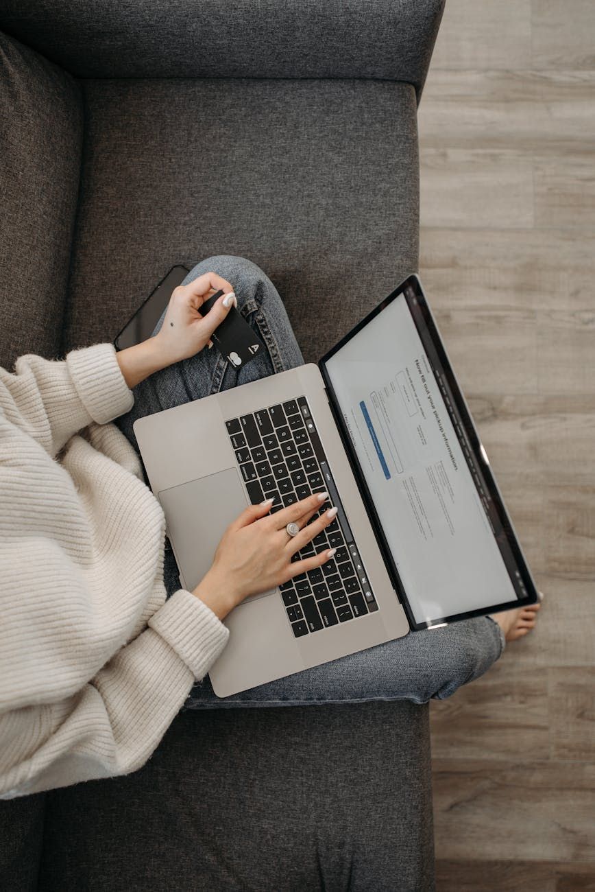 Person on a dark sofa typing on a silver laptop, viewed from above.