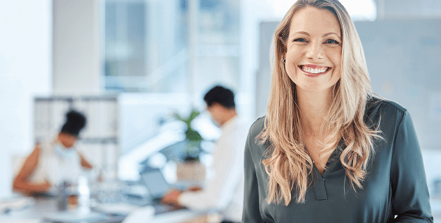 Professional blonde woman smiling confidently in a modern office, colleagues working behind.