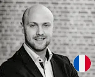 Smiling bald man with a beard in a black and white photo, French flag in corner.