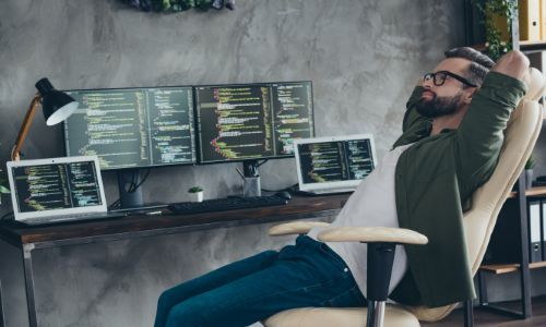 A bearded man relaxes in a chair, contemplating code on multiple computer monitors.