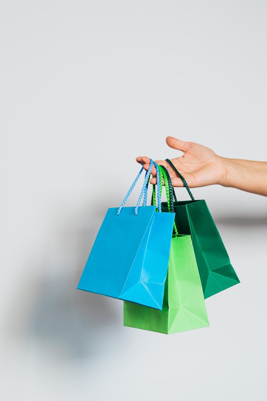 A person's hand holds three colorful shopping bags: blue, light green, and dark green.